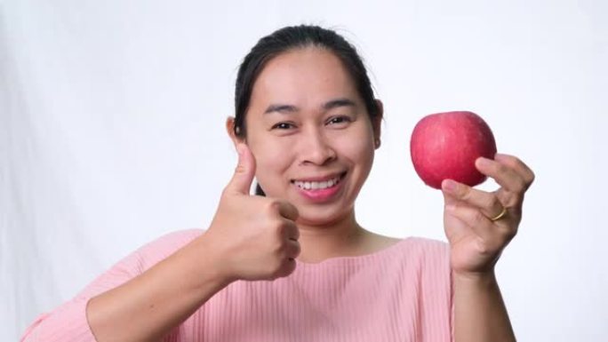 亚洲女子手持红苹果，竖起大拇指，微笑着在工作室的白色背景上展示健康的牙齿