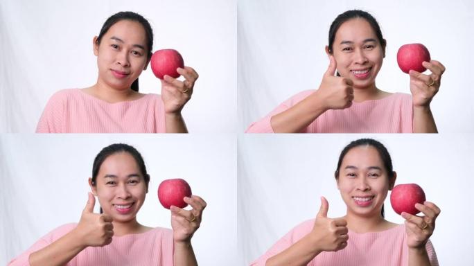 亚洲女子手持红苹果，竖起大拇指，微笑着在工作室的白色背景上展示健康的牙齿