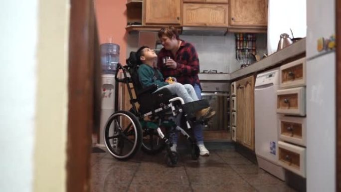 母亲带着轮椅上的残疾儿子在家做饭，同时给他一杯水-有残疾孩子的家庭
