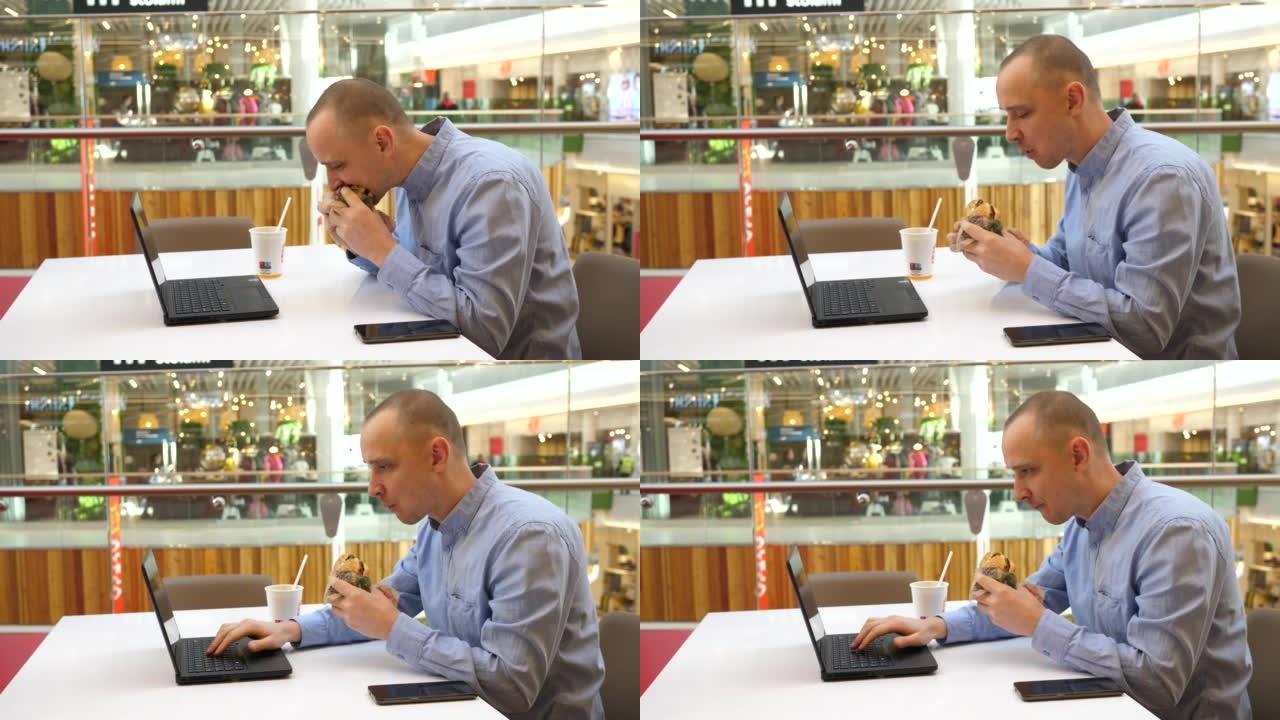 年轻人在城市购物中心美食广场远程工作，吃着美味的大汉堡咀嚼和打字笔记本电脑