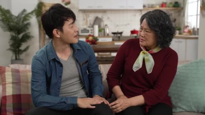 亚洲成年儿子的慢动作在家里用手势向母亲解释他的工作，因为他们坐在沙发上很近。成熟的女人听着点头