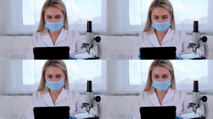 女研究化学家科学家戴着防护口罩和手套，穿着医疗服，带着平板电脑和显微镜，在实验室进行研究