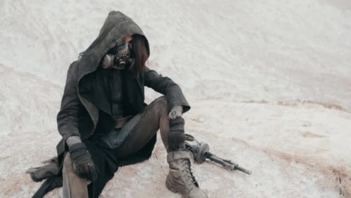 手持慢镜头后世界末日的女人穿着邋遢的衣服戴着黑色的斗篷保护防毒面具呼吸系统坐在沙漠中的白山上。赛博朋