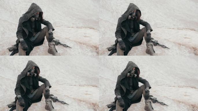 手持慢镜头后世界末日的女人穿着邋遢的衣服戴着黑色的斗篷保护防毒面具呼吸系统坐在沙漠中的白山上。赛博朋
