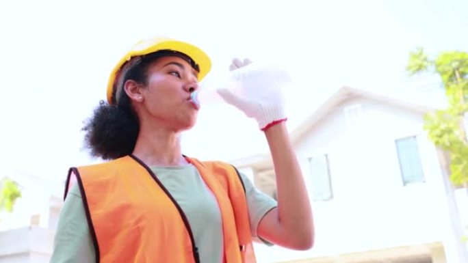 特写脸黑人女人。戴着黄色安全帽头盔的非裔美国女工程师工人在建筑工地上喝水。