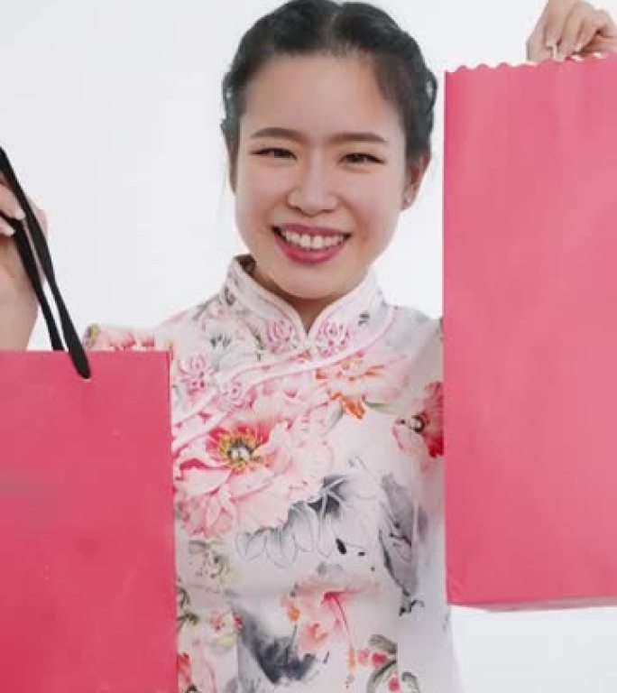 中国/亚洲女性在春节期间双手拿着红色和粉红色的购物袋，白色背景，穿着旗袍白色碎花图案连衣裙