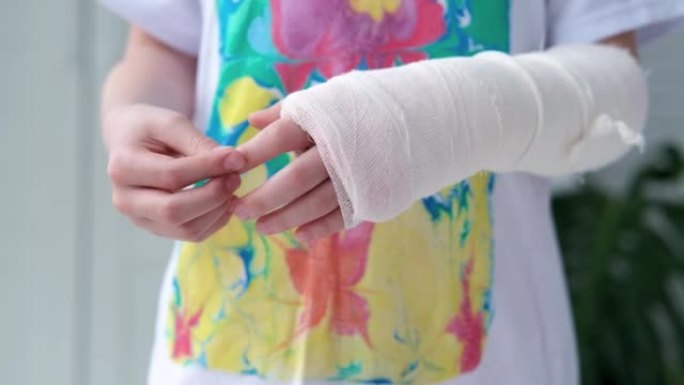 用白色石膏绷带包裹的断手手指做按摩的孩子。医疗保健概念。女孩在多色t恤的背景下折断了手臂