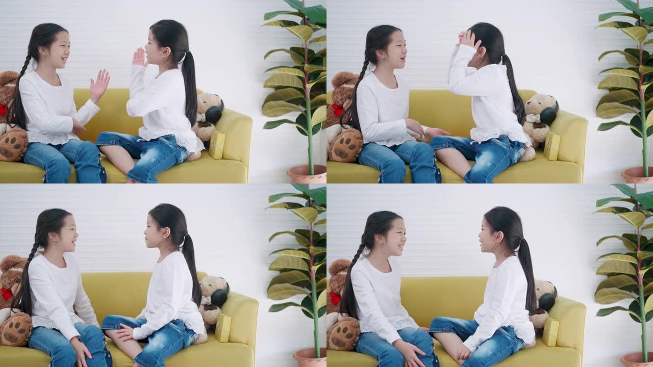 两个小学的亚洲女孩坐在家里的沙发上一起唱歌。混合年龄的孩子或女孩朋友是白色t恤在课前一起唱歌，并兴奋