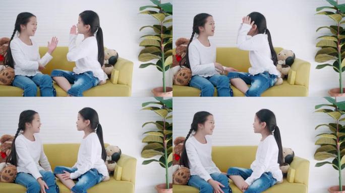 两个小学的亚洲女孩坐在家里的沙发上一起唱歌。混合年龄的孩子或女孩朋友是白色t恤在课前一起唱歌，并兴奋