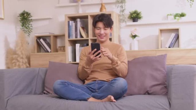 坐在房子的沙发上，一个亚洲人在他的智能手机上工作。男人在使用社交媒体应用程序时工作。在家工作的概念。