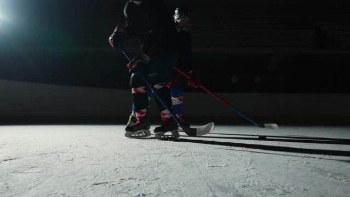 两名运动员曲棍球运动员在冰场上滑行并运球，用棍子击中冰球。穿着制服，头盔和棍棒的年轻人在聚光灯下的黑