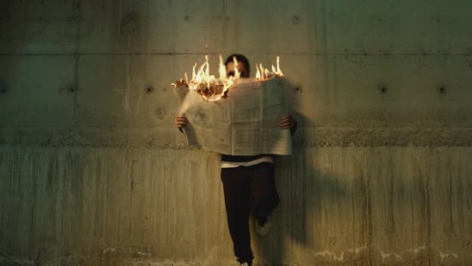 男性艺术家的镜头，戴着墨镜站在墙边，在燃烧期间拿着和阅读报纸。虚假或禁止新闻的概念或象征。男人读燃烧