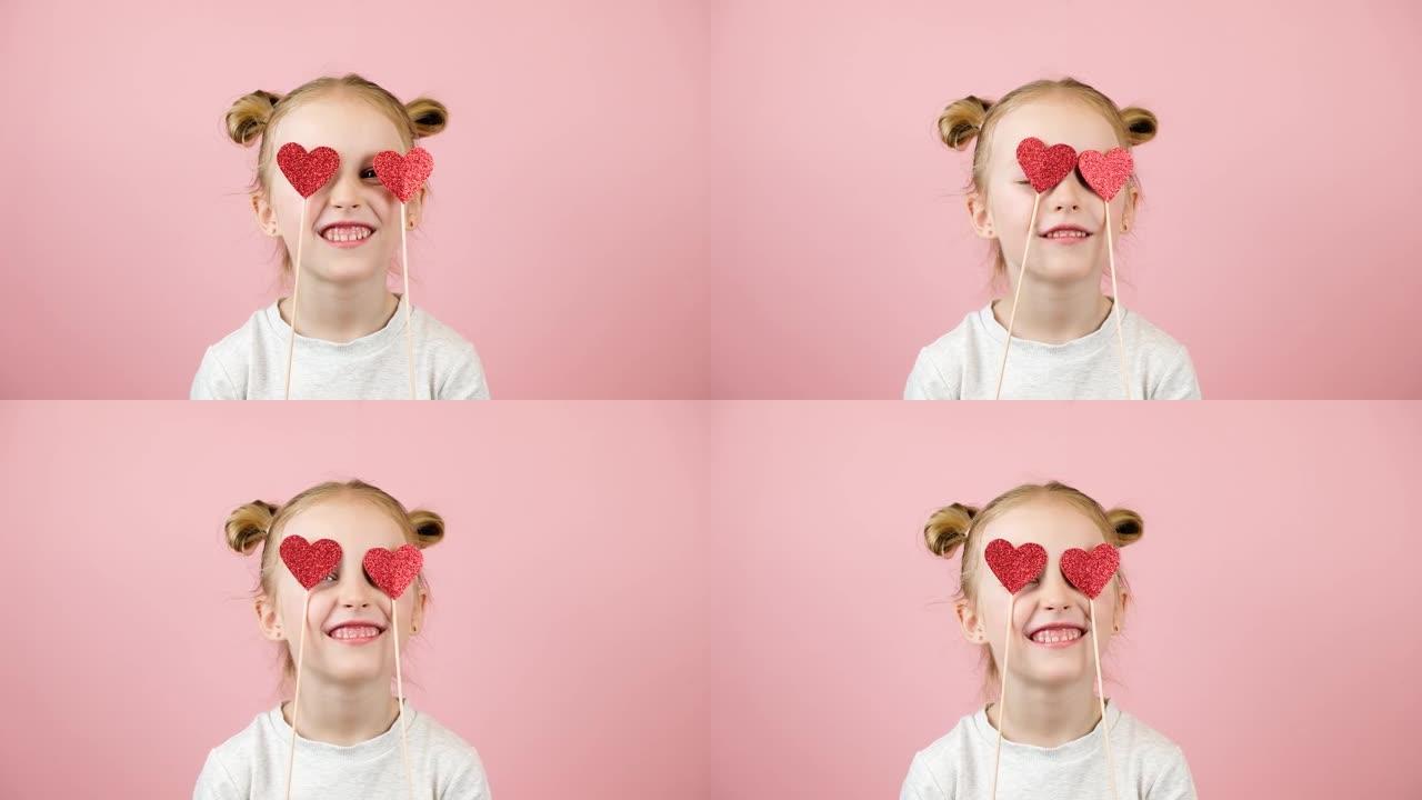 有趣的金发小女孩微笑着玩粉色背景的红心玩具。情人节或母亲节概念