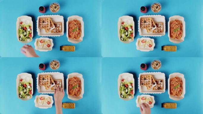 送餐俯视图，在蓝色背景上的一次性容器中带走餐点。午餐盒配有煮熟的蔬菜沙拉和意大利面。将蓝莓放在比利时