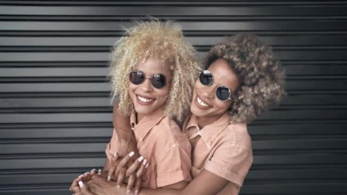 年轻的黑人双胞胎姐妹的肖像，金色的头发和墨镜，微笑着，用爱和亲情拥抱着彼此