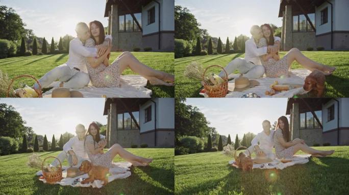 浪漫的野餐男孩和女孩坐在美丽的房子附近的草坪上，快乐地拥抱着玩小狗