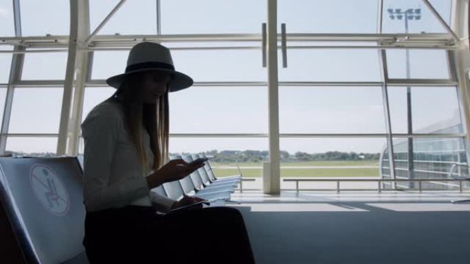 年轻女性旅行者，穿着时尚，休闲或商务，坐在一个大型国际机场枢纽的玻璃窗旁，手里拿着手机，滚动着智能手