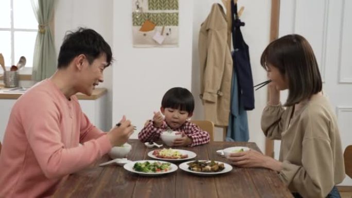 快乐的亚洲父亲、母亲和学龄前男孩在家一起吃午饭。这对恩爱的夫妇互相把食物和儿子的碗放在一起