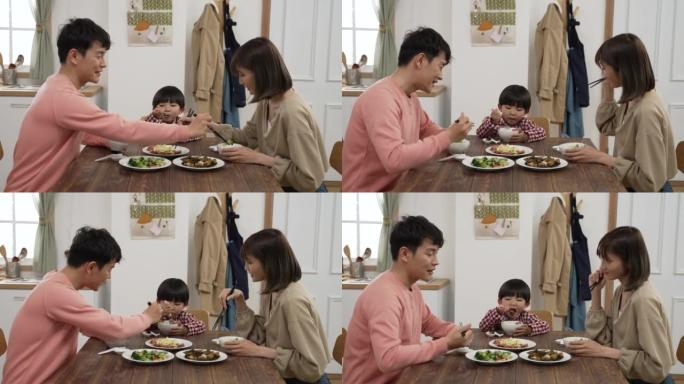 快乐的亚洲父亲、母亲和学龄前男孩在家一起吃午饭。这对恩爱的夫妇互相把食物和儿子的碗放在一起