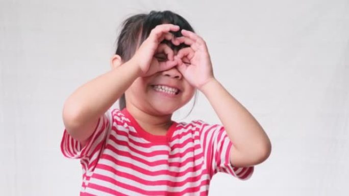 可爱的小女孩戴着指法眼镜做着滑稽的脸，形状就像用双筒望远镜在白色工作室背景上看一样。
