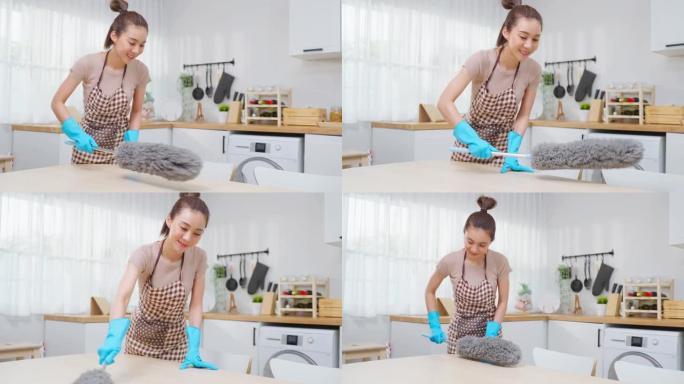 亚洲清洁服务女工在家厨房打扫卫生。漂亮的年轻管家清洁工穿围裙，用鸡毛掸子擦拭餐桌做家务或家务。