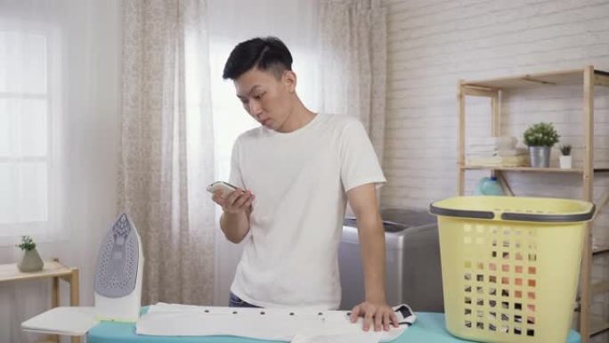 亚洲年轻人从口袋里掏出手机查看信息，在家里明亮的洗衣房里熨衣服时，高兴地笑着。