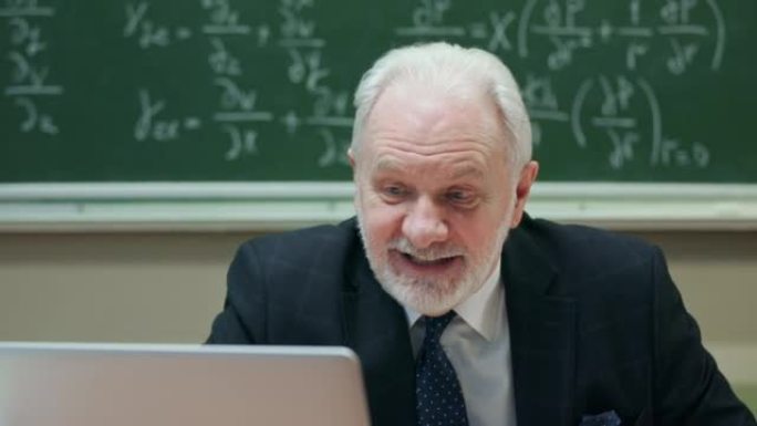 高级白发老师教授与学生交谈，在大学或大学教室上课时使用笔记本电脑解释数学公式，背景黑板。教育和学习理