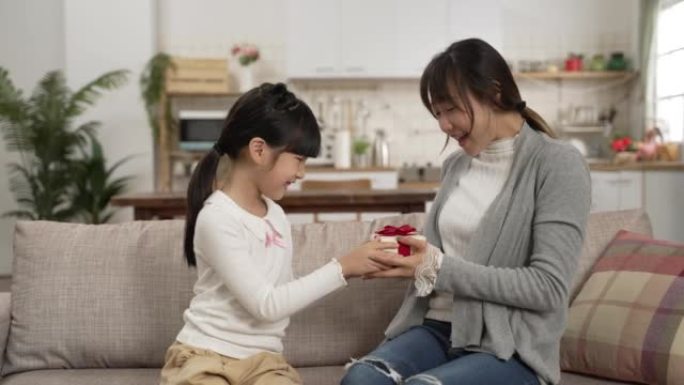 惊讶的亚洲母亲拥抱女儿在家中获得母亲节礼物的慢动作。当妈妈遮住眼睛时，女孩从背后展示礼物