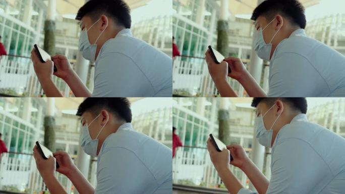 亚洲男孩戴着口罩，坐在百货商店玩手机游戏，新常态和社交疏远的生活方式概念。