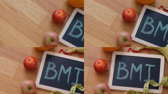 黑板上的缩写BMI (身体质量指数) 和一些水果，它周围有一个水瓶和卷尺。