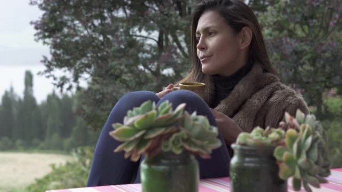 美丽的拉丁女人坐在小屋外面喝咖啡，看着雨