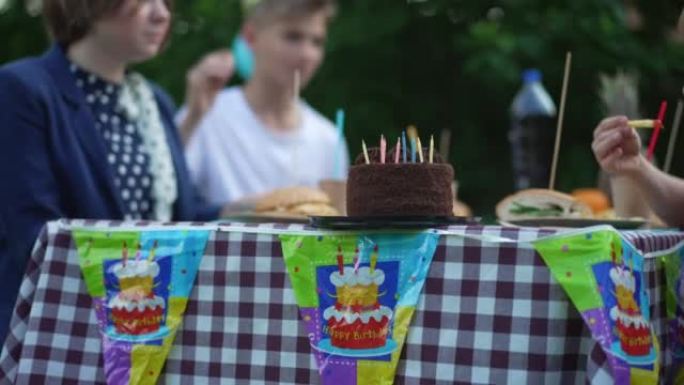 后院桌子上的巧克力生日蛋糕，一群快乐的模糊孩子坐在背景下慢动作大笑。放松的高加索朋友在户外庆祝。