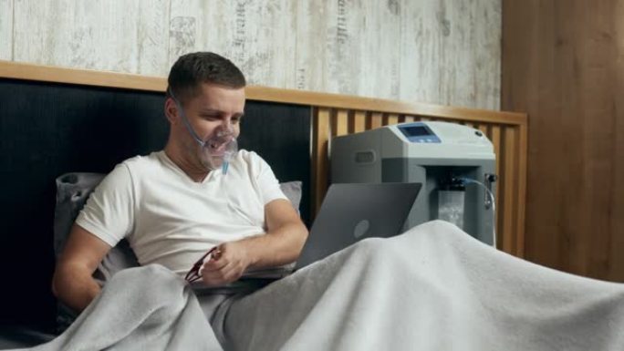 一个生病的年轻人的肖像使用氧气面罩和氧气浓缩器在家中治疗并在笔记本电脑上工作。