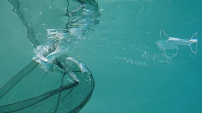 医用口罩在蓝色水柱水下漂移。Net从水中去除口罩，清洁环境并消除对自然的威胁。海洋中的污染问题概念。
