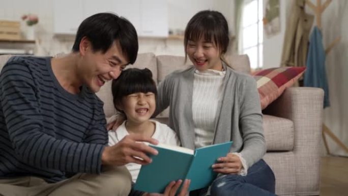 快乐的亚洲父亲和母亲在家给女学生女儿读书的慢动作。当男人讲述书中的有趣部分时，他们都笑了