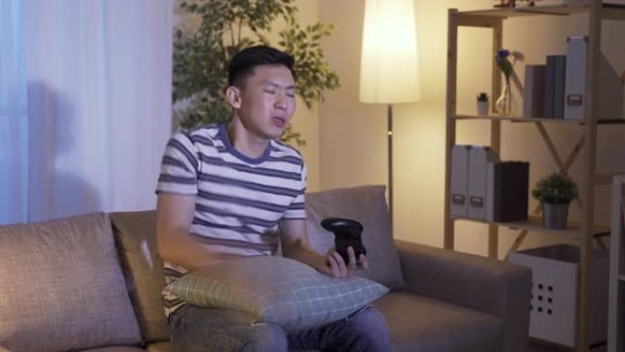 心烦意乱的亚洲男子拍打枕头，双手拿着操纵杆沮丧地抱着头，因为他失去了家里客厅沙发上的电子游戏
