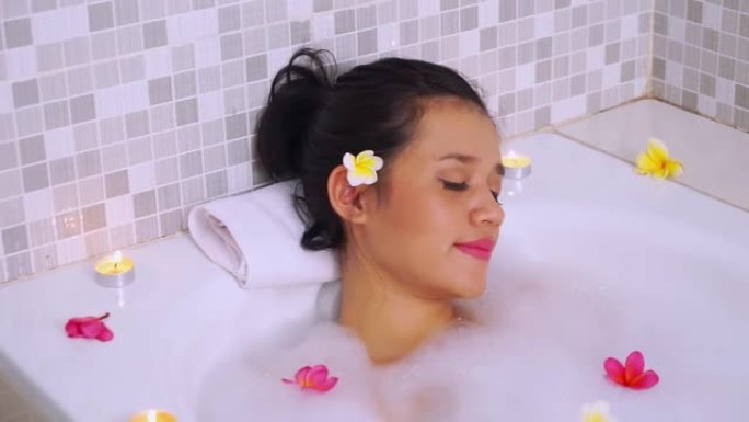 一个年轻美丽的亚洲女人在私人浴缸上放松的视频片段，用肥皂泡、素馨花和蜡烛