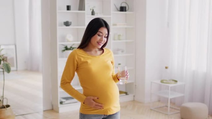 身体水合作用。年轻的亚洲孕妇在家中用玻璃杯喝水