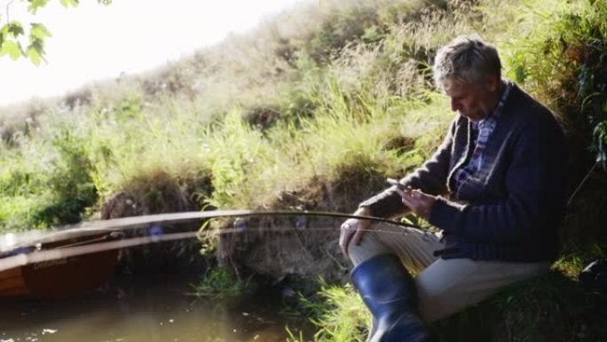 成熟的男人在阳光明媚的夏日河岸钓鱼和使用智能手机
