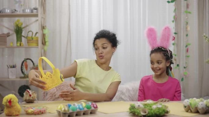 妈妈和女儿用有趣的兔子耳朵装饰鸡蛋篮。非裔美国妇女和小女孩坐在家里装饰精美的房间里的桌子旁。复活节快