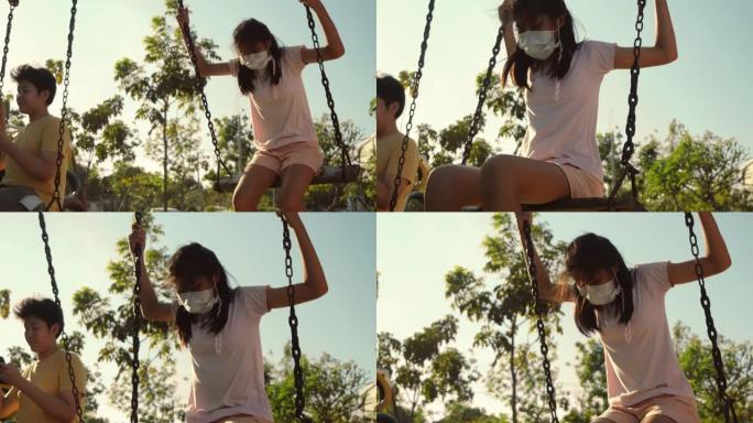 亚洲女孩戴着口罩和她的兄弟在户外操场上玩秋千，带着太阳光，活跃和新的正常生活方式概念。