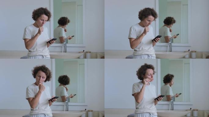 一位穿着家装的漂亮年轻女子用牙刷刷牙，照顾她的口腔，在智能手机上看和写信息。早上在浴室