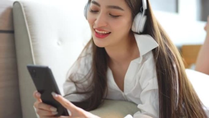美丽的亚洲女人穿着性感的连衣裙享受在客厅沙发上戴着耳机在智能手机技术上听音乐