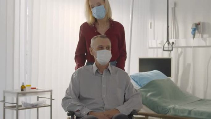 戴防护面罩的高级妻子将出院的男性患者推上轮椅住院