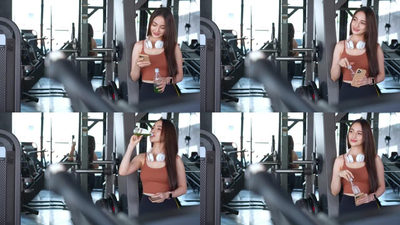 一名亚洲妇女在健身房休息，在健身时间用减肥机锻炼后，用毛巾擦干脸，喝有机蔬菜汁，用智能手机放松。