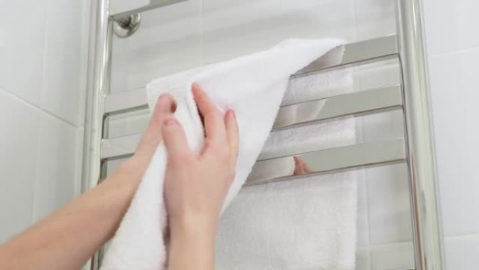 用挂在加热毛巾架上的白色毛圈柔软毛巾擦拭湿手。立式电动毛巾取暖器。干燥器上的方形铬管