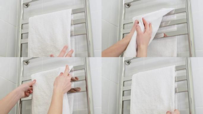 用挂在加热毛巾架上的白色毛圈柔软毛巾擦拭湿手。立式电动毛巾取暖器。干燥器上的方形铬管