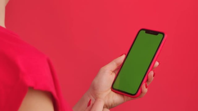 女人拿着红色工作室背景上的绿色工作区模拟屏幕的手机。女性手触摸、点击、滑动。广告区域。模板位置。特写