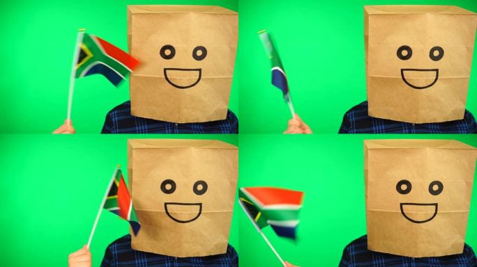 头上戴着纸袋的男子的肖像，在绿色的背景下挥舞着南非国旗，面带微笑。