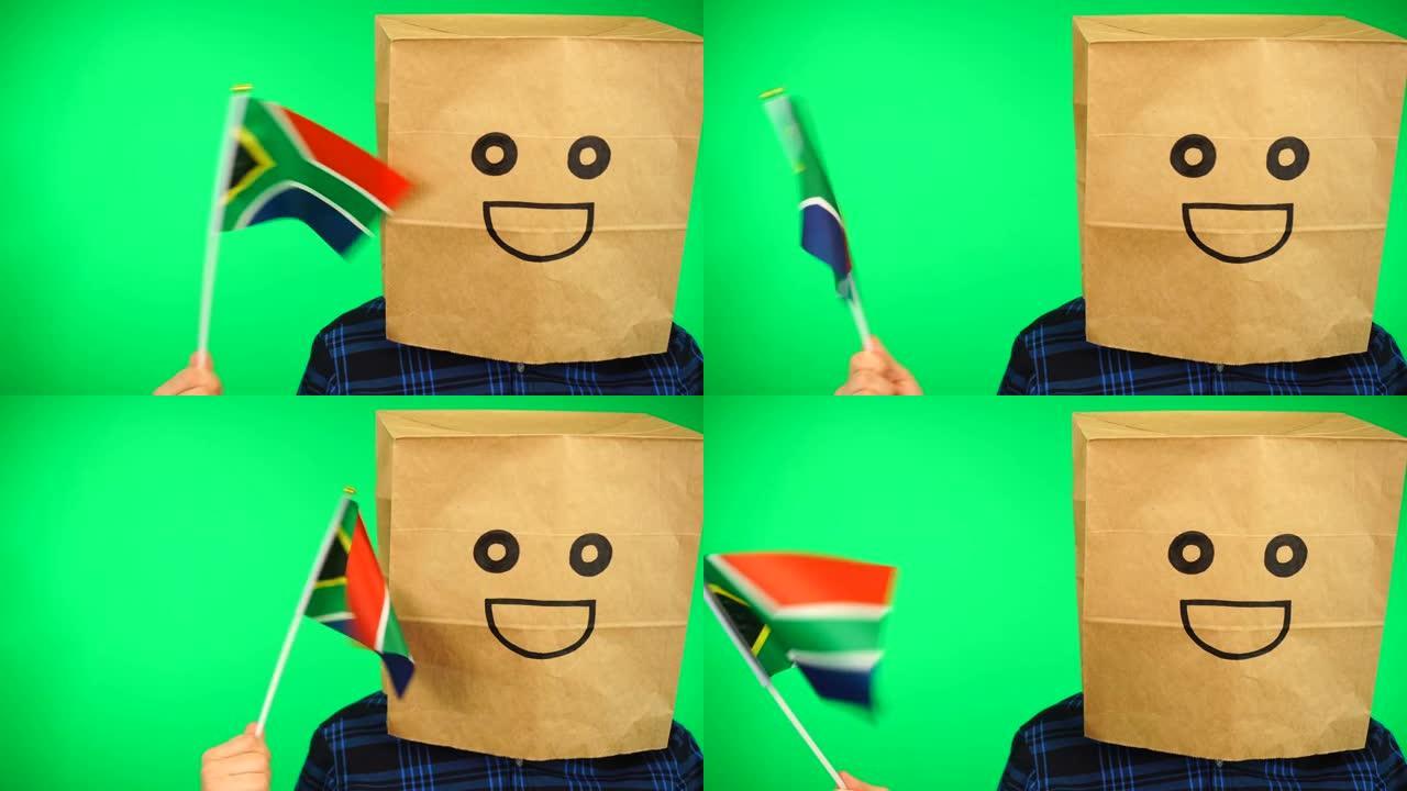 头上戴着纸袋的男子的肖像，在绿色的背景下挥舞着南非国旗，面带微笑。